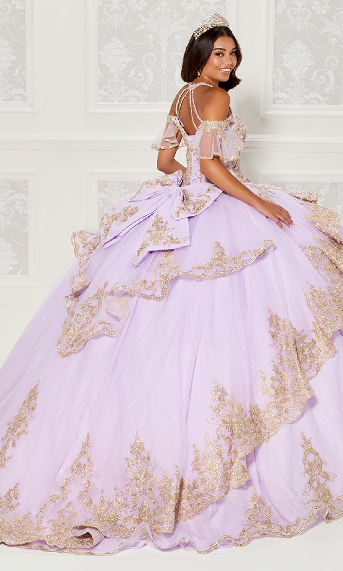 A woman wearing the Quinceanera Dress PR30118 By Princesa with a tiara stands before an ornate white wall, looking over her shoulder and smiling.